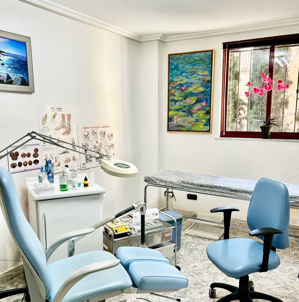 Una luminosa sala de reconocimiento médico con una silla azul para el paciente, un taburete para el médico, una mesa de reconocimiento y equipamiento. Las paredes blancas están decoradas con carteles anatómicos y obras de arte. Una ventana con una orquídea en maceta da la bienvenida a la luz natural al espacio de Ana Iglesias podóloga.