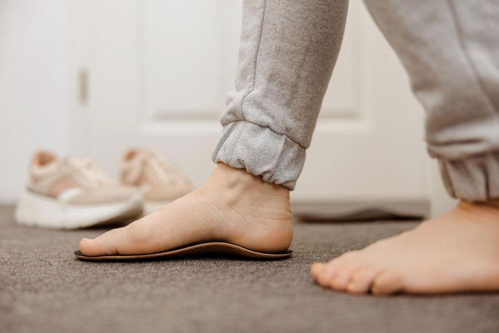 Una persona vestida con un pantalón de chándal gris claro permanece descalza en el interior, colocando un pie sobre una plantilla ortopédica, un método recomendado a menudo por Ana Iglesias podóloga. Al fondo, en el suelo, se ven unas zapatillas deportivas de color beige.