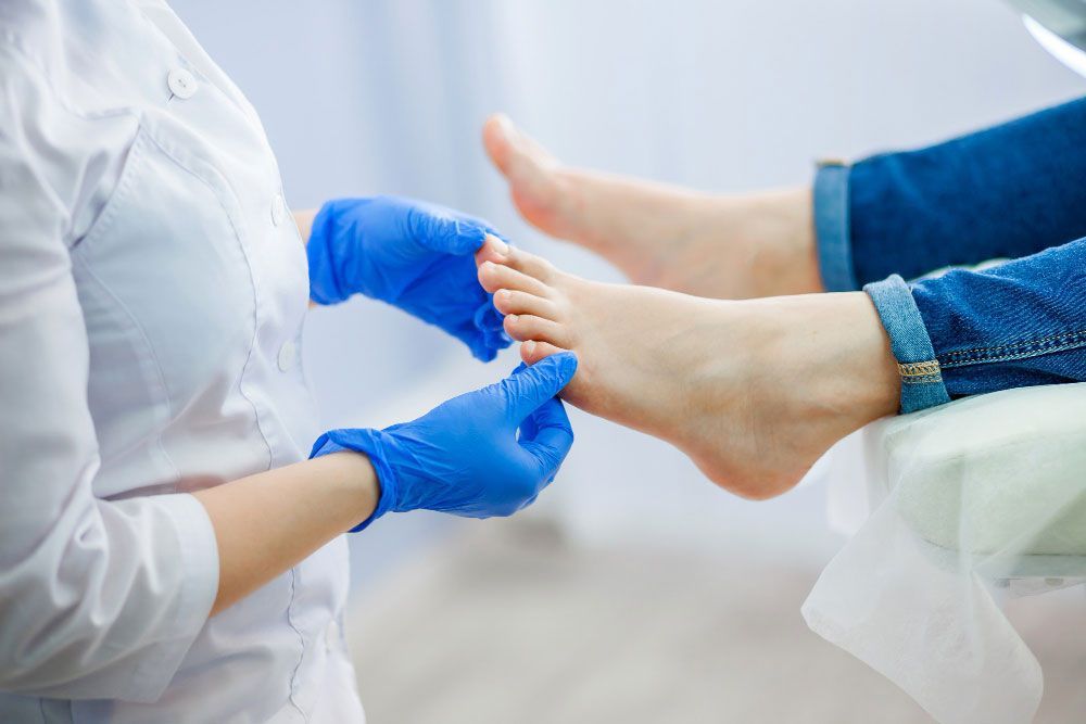 Ana Iglesias podóloga, con guantes médicos azules, examina los pies descalzos de otra persona, que está sentada con los pantalones arremangados, en un entorno clínico.