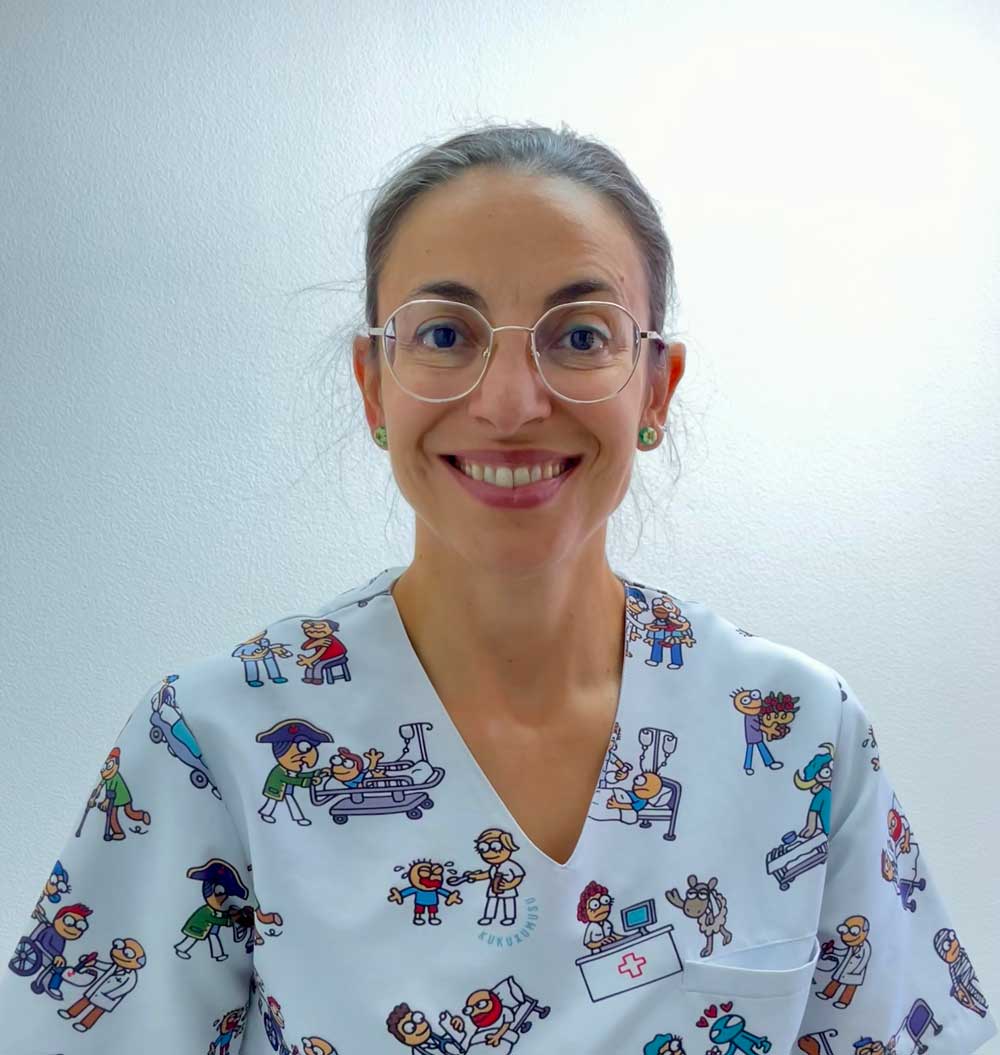 Una mujer sonriente con gafas y una colorida bata médica con ilustraciones sanitarias de dibujos animados frente a un fondo claro liso: Ana Iglesias podóloga.
