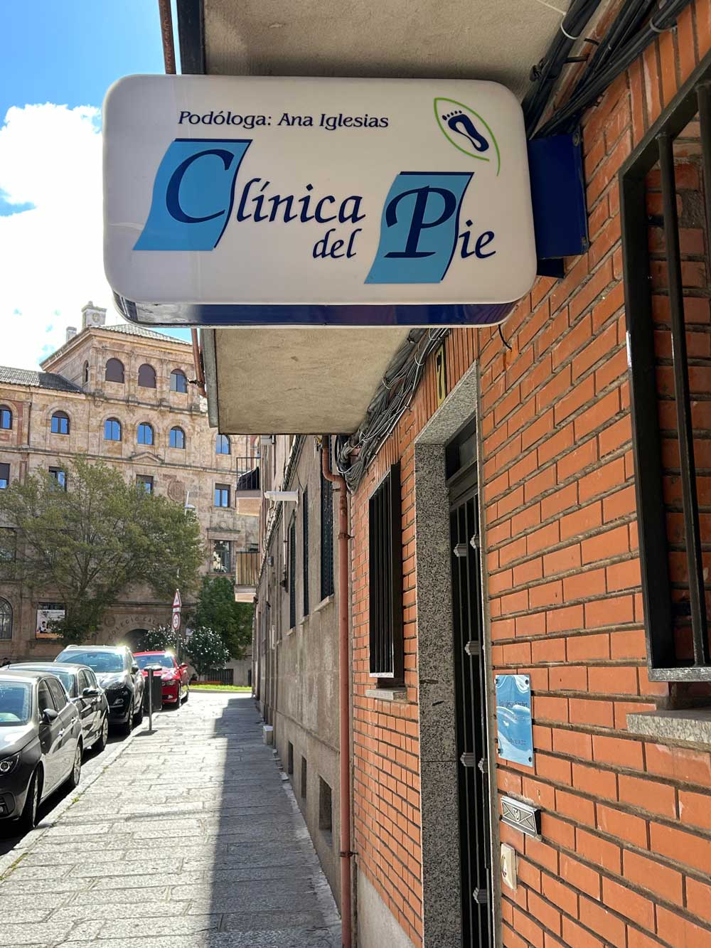 Vista de la calle de un edificio de ladrillo con un cartel de Ana Iglesias podóloga, "Clínica del Pie", sobre la entrada. Varios coches aparcados se alinean en la calle, con un gran edificio visible al fondo.